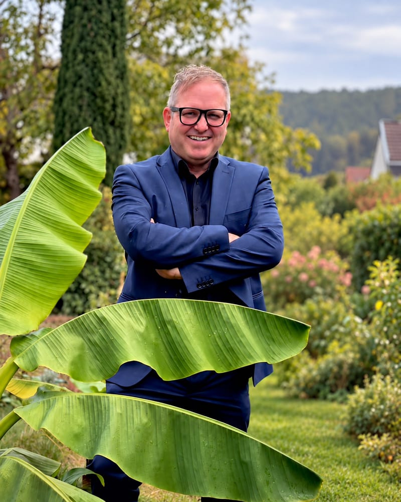 Jörg „Schorni“ Wittmer mit verschränkten Armen im Garten hinter einer Bananenstaude, im Hintergrund Bäume und die bewaldeten Hügel des Dahner Felsenlands